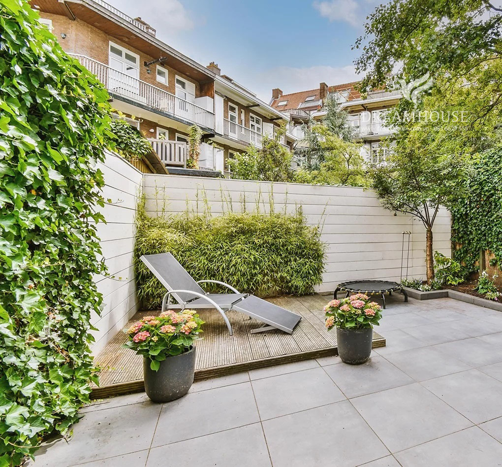 35-y-tuong-thiet-ke-canh-quan-san-vuon-cho-nam-2025-cho-nguoi-dam-me-3 Khu vườn lát đá nhỏ có sàn nâng ở góc, có ghế tắm nắng và cây trồng trong chậu.